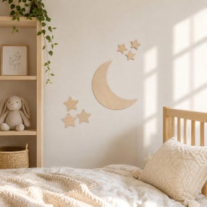 Può includere: Un set di decorazioni da parete in legno di colore chiaro composto da una grande luna crescente e sei stelle a cinque punte di varie dimensioni, disposte in gruppo su una parete liscia sopra un letto con cuscino e biancheria color crema.