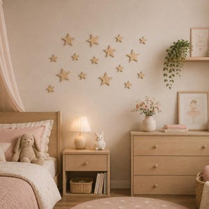 Könnte beinhalten: Eine Ansammlung von hölzernen sternförmigen Wandaufklebern in verschiedenen Größen, angeordnet an einer Wand über einem Bett aus hellem Holz, einem Nachttisch mit Lampe und einer Kommode. Der Raum enthält zartrosa Bettwäsche und einen Stoffhasen.
