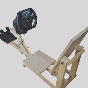 Può includere: Un simulatore di corse in legno con un volante nero con display integrato, un set di pedali e un telaio in legno. Il telaio è costruito in legno chiaro e comprende una struttura simile a un sedile per l'utente.