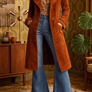 Peut inclure: Un long manteau en daim orange brûlé avec un col à revers, des manches longues et une fermeture boutonnée, porté sur un haut beige à lacets et un jean bleu évasé taille haute. Le manteau présente deux poches latérales et des poignets boutonnés.
