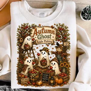 May include: A white crewneck sweatshirt featuring a graphic print of three ghosts and a scarecrow picking apples in an autumn orchard. The text reads Autumn Ghost Apple Picking, Fresh Apples, and Pick Apples Cozy Days Happy Ghosts.