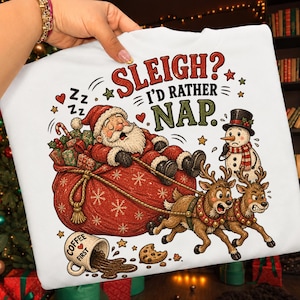 Pode incluir: Uma camisola branca com um gráfico do Pai Natal a dormir num grande saco vermelho de presentes, puxado por duas renas. Um boneco de neve está por perto. O texto diz "SLEIGH? I'D RATHER NAP." Uma caneca entornada com "COFFEE FIRST" e uma bolacha estão em baixo.