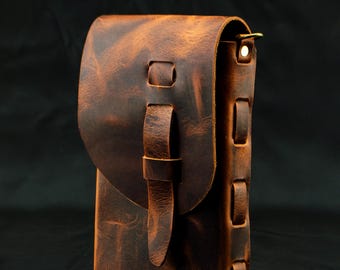 May include: A vertical brown leather pouch with a distressed finish, featuring a fold-over flap secured by a leather strap and loop closure. The side panel is constructed with visible leather lacing details and a small metallic hardware ring.