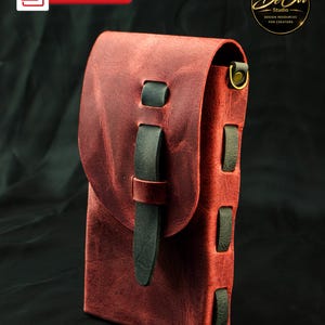 May include: A vertical rectangular pouch crafted from distressed red leather, featuring a fold-over flap secured by a dark green leather strap. The sides are laced with dark green leather strips and include a brass-toned metal ring attachment.