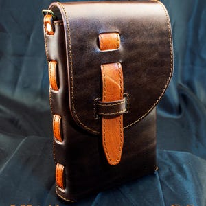 May include: A dark brown leather bag featuring a lighter tan leather strap closure and side accents. The bag has a flap top secured by a vertical strap. Text at the bottom reads Full Tutorial & PDF Download and De Ovi Studio.