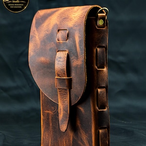 May include: A vertical brown leather pouch with a distressed texture, featuring a rounded flap closure secured by a leather strap loop. The side panel displays a series of rectangular cutouts with woven leather detailing and a brass metal D-ring.