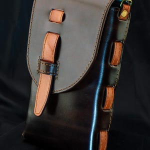 May include: A dark brown leather pouch featuring a lighter tan leather strap closure and side accents. The pouch has visible stitching along the edges and a brass-colored metal ring at the top. Text at the bottom reads Full Tutorial & PDF Download De Ovi Studio.