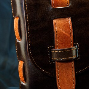 May include: A dark brown leather bag featuring a contrasting tan leather strap closure and side lacing details. The bag is constructed with visible stitching and includes a vertical strap secured by a loop. The text De Ovi Studio is visible.