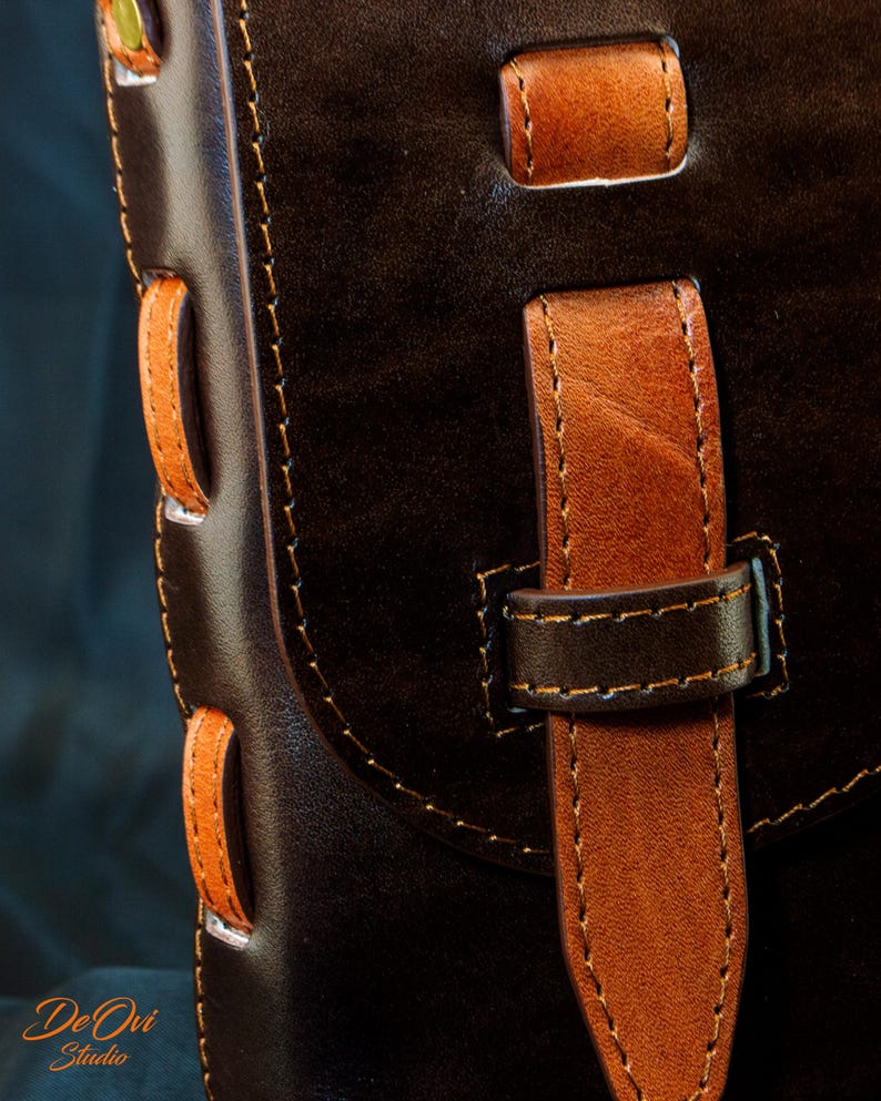 May include: A dark brown leather bag featuring a contrasting tan leather strap closure and side lacing details. The bag is constructed with visible stitching and includes a vertical strap secured by a loop. The text De Ovi Studio is visible.