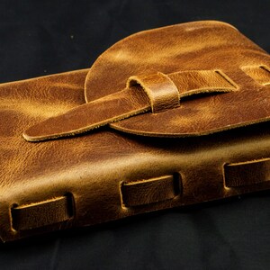 May include: A rectangular brown leather pouch featuring a textured surface, a rounded flap closure secured with a leather strap, and a series of rectangular cutouts along the side edge with a small metal ring attached to the corner.