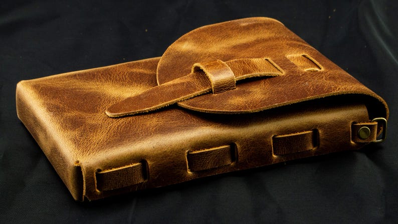 May include: A rectangular brown leather pouch featuring a textured surface, a rounded flap closure secured with a leather strap, and a series of rectangular cutouts along the side edge with a small metal ring attached to the corner.