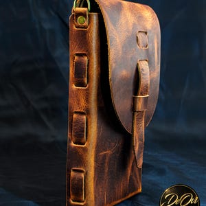 May include: A brown leather pouch with a textured surface, featuring a flap closure secured by a vertical leather strap. The side of the pouch displays three rectangular leather loops and a brass D-ring attachment at the top corner.