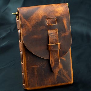 May include: A vertical brown leather pouch with a distressed finish, featuring a rounded flap closure secured by a leather strap and loop. The sides are constructed with visible leather lacing details and brass hardware attachments at the top corners.