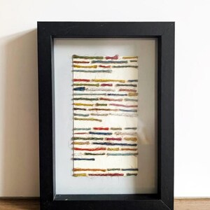 May include: A framed artwork featuring a rectangular piece with colorful horizontal lines. The lines are in shades of red, blue, yellow, and green, set against a white background. The frame is black.