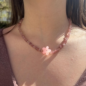 Peut inclure: Un collier de perles roses avec un pendentif en forme de fleur. Le collier est composé de petites perles roses irrégulières et d'un pendentif en forme de fleur rose clair au centre. Le collier est porté autour du cou.