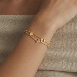 Peut inclure: Un bracelet doré à deux rangées, orné de breloques en forme de trèfle et d'étoile. Une bague délicate avec de petites pierres orne un doigt. Le bijou est porté sur un bras, sur un fond neutre.