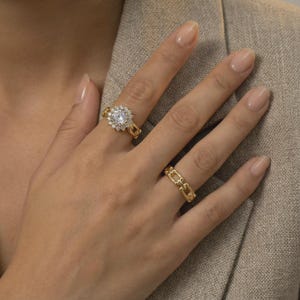 Peut inclure: Deux bagues en or sont présentées sur une main. L'une comporte une grosse pierre précieuse transparente entourée de petites pierres, sertie sur un anneau à maillons de chaîne. L'autre est un design simple à maillons de chaîne. La main est contre une veste de couleur neutre.