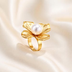 Op de afbeelding: Een goudkleurige ring met een bloemvormig ontwerp en een grote, witte parel in het midden. De bloemblaadjes hebben een getextureerde afwerking. De ring ligt op een zachte, crèmekleurige achtergrond.