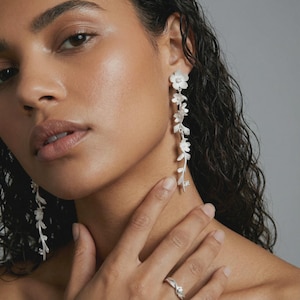Peut inclure: Boucles d'oreilles pendantes argentées avec un motif floral, composées de petites fleurs et de feuilles. Les boucles d'oreilles sont ornées de détails scintillants. Une bague argentée assortie avec un motif floral est également visible.
