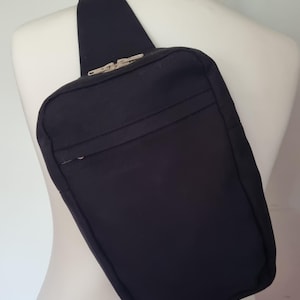 May include: A solid black crossbody sling bag featuring a single wide shoulder strap, a rectangular main compartment with a horizontal zippered front pocket, and silver-toned metal zipper pulls, worn across the torso on a white mannequin.