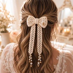 May include: A cream-colored, crocheted hair bow with a pearl-beaded center and dangling lace ribbons. The ribbons are adorned with pearls and gold-colored floral accents. The bow is set against a backdrop of wavy brown hair.