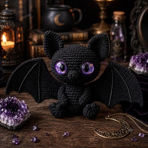 Könnte beinhalten: Eine handgefertigte schwarze Häkel-Fledermaus mit großen, lila und goldfarben gefleckten Augen und ausgebreiteten Flügeln. Die Fledermaus sitzt auf einer Holzoberfläche, umgeben von Amethystkristallen und einer Mondhalskette. Der Hintergrund umfasst Kerzen und antike Bücher.