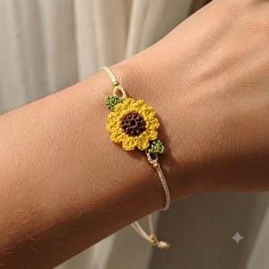 May include: A delicate bracelet featuring a small, crocheted sunflower motif with bright yellow petals and a dark brown center. The flower is accented by two tiny green crocheted leaves and is attached to a thin, off-white cord with small gold beads.