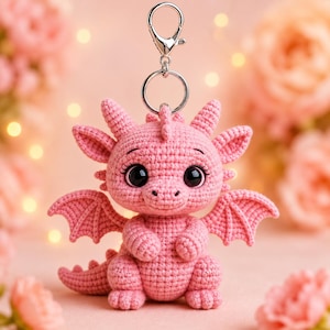 Pode incluir: Um chaveiro de dragão rosa de crochê com grandes olhos pretos, pequenas asas e um chaveiro prateado. O dragão está sentado, com os braços e pernas visíveis. O fundo é rosa suave com flores desfocadas.
