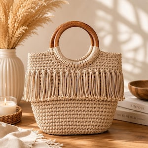 Peut inclure: Un sac à main en crochet beige avec des poignées en bois. Le sac présente une frange sur le bord supérieur. Les poignées sont circulaires et en bois brun clair. Le sac est posé sur une surface en bois.