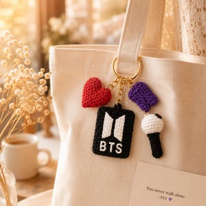 May include: A cream tote bag with a gold keyring holding four crochet charms. The charms include a red heart, a black rectangle with white lettering and logo, a purple shape, and a white and black microphone. A card with text is visible.