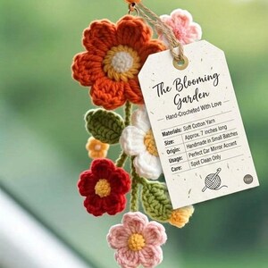 May include: A hand-crocheted car mirror accent with a floral design in orange, red, pink, yellow, and white. The flowers are attached to a string with a tag that reads "The Blooming Garden". The accent is approximately 18 cm long.