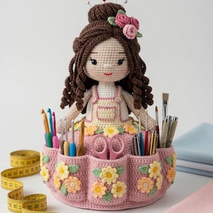 May include: A handmade crochet doll with brown hair and a floral crown, holding a pink organiser. The organiser features pockets filled with crochet hooks, scissors, and pens. A yellow measuring tape is visible.