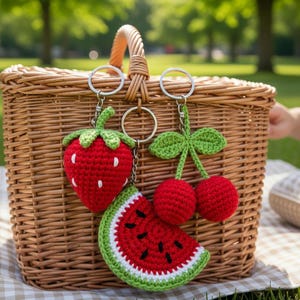 Puede incluir: Una cesta de picnic de mimbre con tres llaveros de frutas de ganchillo: una fresa roja, una rodaja de sandía y un par de cerezas. Los llaveros están sujetos a anillos plateados. El fondo es un parque con hierba.