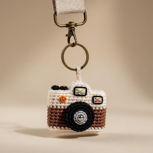 Op de afbeelding: Een gehaakte camera sleutelhanger met een bruin en wit lichaam, zwarte lens en een metalen ring en karabijnhaak. De sleutelhanger heeft een beige band en hangt tegen een neutrale achtergrond. De camera heeft kleine details in zwart, oranje en geel.