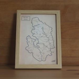 May include: A framed map of The Peak District printed on off-white paper with black ink lines and text. The map features geographical outlines, labeled locations, and dashed paths. The frame is made of light-colored wood with a visible grain pattern.