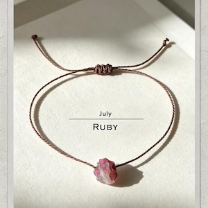 Pode incluir: Uma pulseira de cordão castanho com uma única pedra preciosa de rubi em bruto de cor rosa-avermelhada no centro. A pulseira tem um fecho de nó deslizante ajustável. O texto July e RUBY está impresso a preto sobre a pedra na superfície.