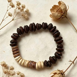 Puede incluir: Un brazalete hecho a mano con cuentas oscuras en forma de frijol y una sección de cuentas redondas de color claro. El brazalete se exhibe sobre un fondo neutro con flores secas, mostrando una estética natural y terrosa. Este accesorio es perfecto para un estilo bohemio.