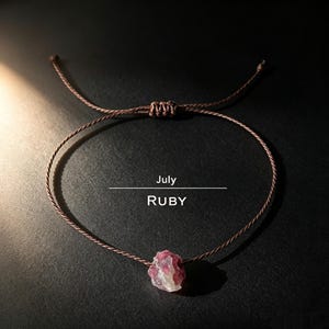 Puede incluir: Una pulsera de cordón marrón con una piedra preciosa de rubí en bruto de color rosa rojizo en el centro. La pulsera tiene un cierre deslizante ajustable. El texto "July" y "RUBY" está impreso en blanco sobre la piedra en el fondo.
