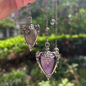 May include: Two teardrop-shaped pendants hanging from silver-toned metal chains. One pendant features a grey stone, the other a purple stone. Both are encased in ornate silver-toned metal frames with intricate filigree, small pentagrams, and wing-like details at the top. Each chain includes small, clear, spherical beads.