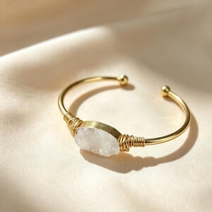 Könnte beinhalten: Ein goldfarbenes, offenes Armband mit einem mittig platzierten, rau geschnittenen weißen Druzy-Stein. Der Stein ist beidseitig mit goldenem Draht umwickelt und mit einem glatten, polierten Metallband verbunden, das in zwei kleinen, abgerundeten Enden ausläuft.