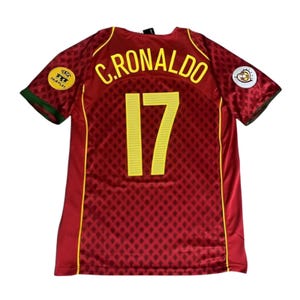 Może przedstawiać: Bordowa koszulka piłkarska z żółtymi akcentami, z napisem "C. RONALDO" i numerem "17" w kolorze żółtym. Koszulka ma wzór w romby i zielone wykończenia na rękawach. Zawiera naszywkę "Fair Play".