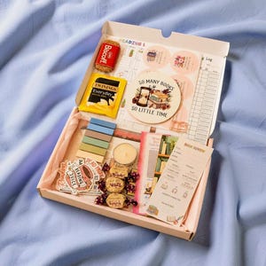 May include: A pink box containing reading-themed items such as a Biscoff biscuit, a tea bag, stickers, a candle, and a book review card. The box also features a coaster with the text "SO MANY BOOKS SO LITTLE TIME".