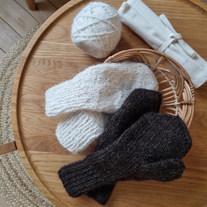 May include: A pair of white knitted mittens and a pair of dark brown knitted mittens are displayed on a wooden tray. A ball of white yarn and a small woven basket are also on the tray. A rolled-up, off-white fabric is tied with a string.