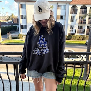 Puede incluir: Una sudadera negra con el logo de los Dodgers de Los Ángeles en pedrería azul y blanca. La persona también lleva una gorra de béisbol color crema con el logo de los Dodgers en negro.