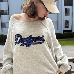 May include: Light gray sweater with the word "Dodgers" in blue script across the front. The sweater has a crew neck and long sleeves. A beige baseball cap with a black "LA" logo is worn.