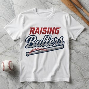 May include: A white short-sleeve t-shirt featuring the text "RAISING Ballers" in red and dark blue with a silver baseball bat graphic underneath. The bat includes a red stitched baseball seam detail across the center of the barrel.