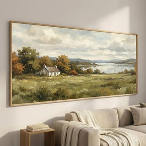 May include: A framed landscape painting depicts a serene lakeside scene. A small white cottage with a metal roof sits amidst green and gold foliage. The artwork features a cloudy sky, calm water, and a wooden frame.