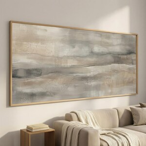 Op de afbeelding: Abstract landschapsschilderij in neutrale tinten. Het kunstwerk toont horizontale banden in beige, grijs en bruin, met getextureerde penseelstreken. Het schilderij is ingelijst in een licht houten frame en hangt boven een beige bank.