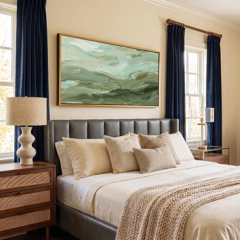 May include: A bedroom scene featuring a bed with a gray leather headboard, cream-colored bedding, and decorative pillows. A large abstract painting in shades of green and gold hangs above the bed. Navy blue curtains frame the windows.