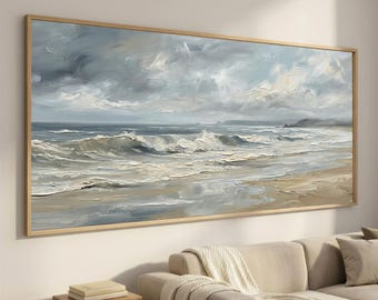 Può includere: Dipinto incorniciato di un paesaggio costiero con onde che si infrangono su una spiaggia sotto un cielo nuvoloso. L'opera d'arte utilizza blu, grigi e beige, con pennellate visibili. Il dipinto è appeso sopra un divano beige e un piccolo tavolino di legno.